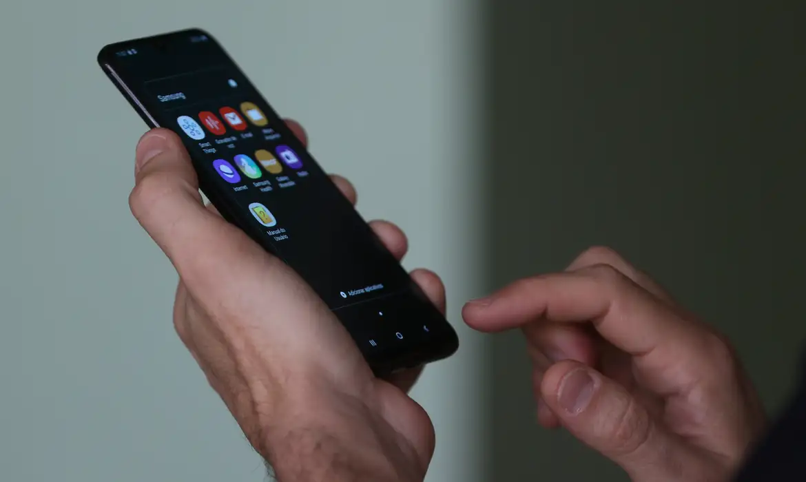 Imagem de uma mão segurando o celular, com vários aplicativos na tela.