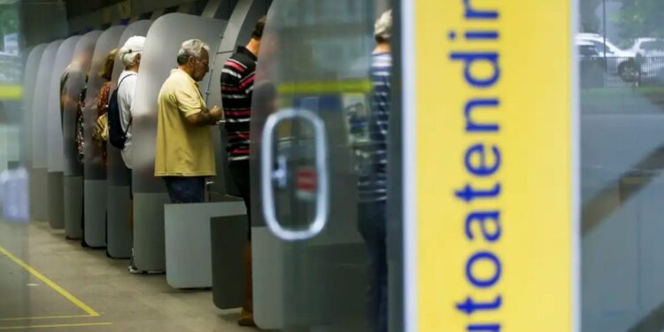 Imagem de pessoas utilizando caixas eletrônicos no banco, ilustrando o funcionamento das agências bancárias durante o carnaval.