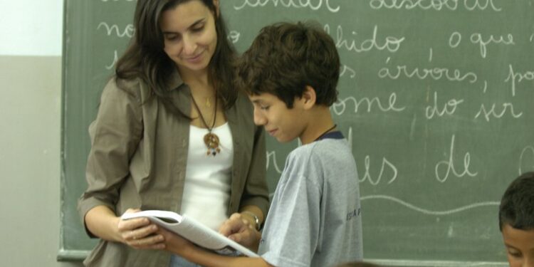 Professor(a) ensina para aluno em sala de aula, ambos olhando para o livro, com quadro negro ao fundo.