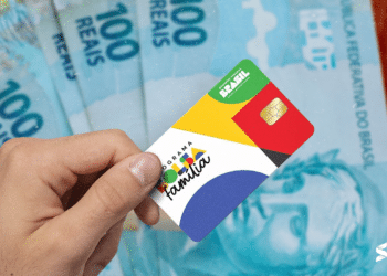 Cartão do Bolsa Família sendo segurado por uma mão, com notas de 100 reais ao fundo, representando o benefício social oferecido pelo governo.