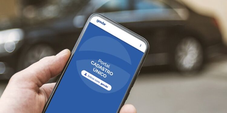 Imagem de um celular mostrando a tela do Portal Cadastro Único no site gov.br, com um carro desfocado ao fundo.