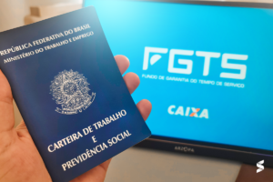 Carteira de Trabalho sendo segurada por uma mão, com a tela ao fundo exibindo o logo do FGTS e da Caixa Econômica Federal.