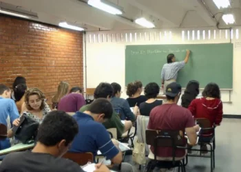 Estudantes em sala de aula assistindo a uma aula, com professor escrevendo no quadro negro.