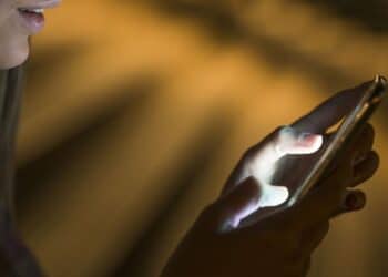 Pessoa usando o celular à noite, com luz de tela iluminando a tela, simbolizando a atenção necessária para evitar golpes.