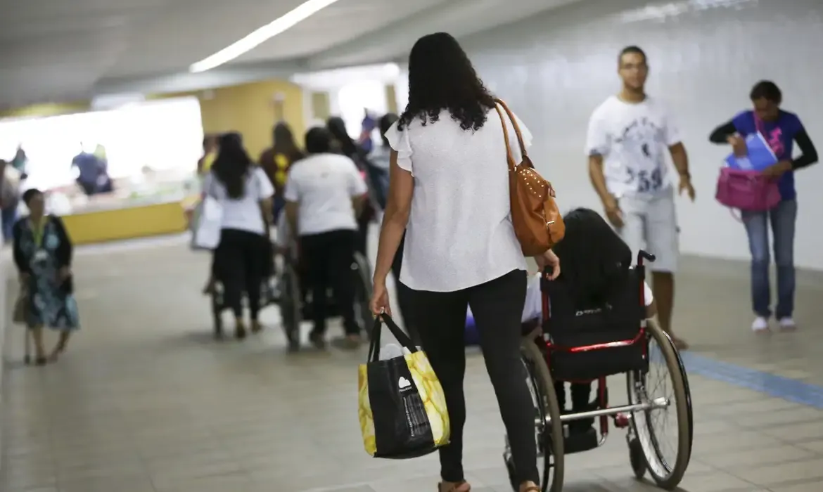 Pessoas caminhando em um espaço público, incluindo uma mulher empurrando uma cadeira de rodas, representando beneficiários do BPC.