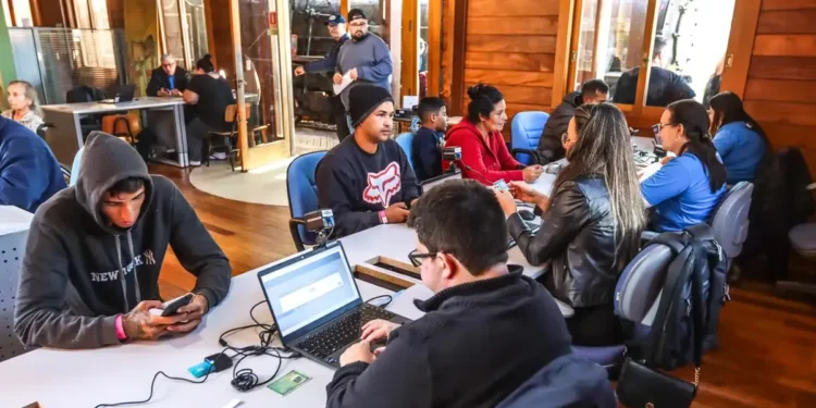 Cidadãos sendo atendidos em um posto de cadastramento do Auxílio Reconstrução, com computadores e documentos sobre a mesa.