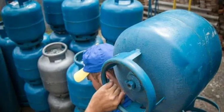Homem carregando um botijão de gás em um depósito, com outros botijões ao fundo.