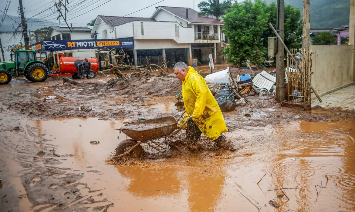 Pessoa com capa de chuva trabalhando em área alagada após enchente, simbolizando os esforços de auxílio em municípios afetados