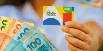 Cartão do Bolsa Família sendo segurado por uma mão, com notas de dinheiro ao fundo, representando o pagamento do benefício.
