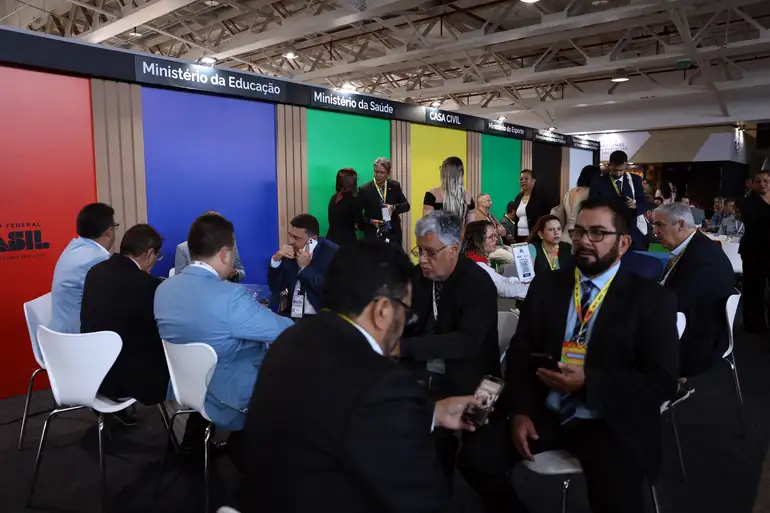 Participantes do Encontro de Novos Prefeitos e Prefeitas em uma mesa de discussão, com stands ao fundo.