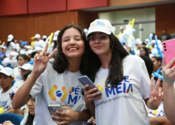 Duas estudantes sorridentes fazem pose em evento do programa Pé-de-Meia, rodeadas por outros participantes.