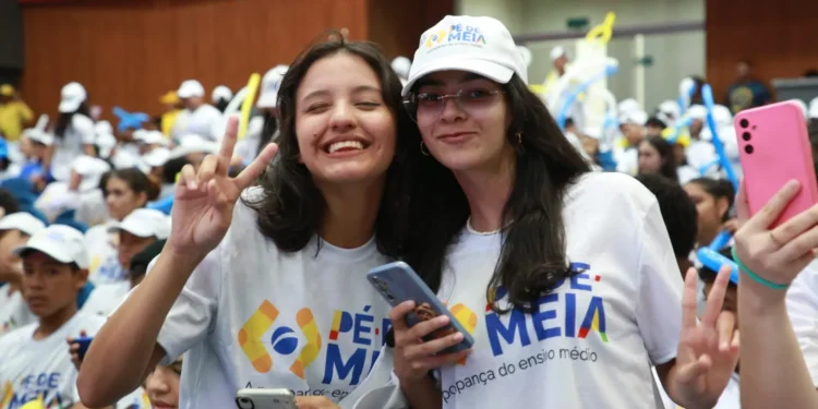Duas estudantes sorridentes fazem pose em evento do programa Pé-de-Meia, rodeadas por outros participantes.