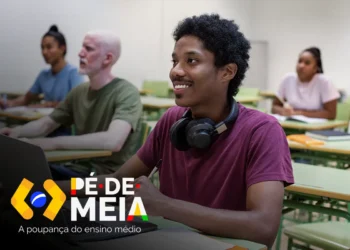 Jovem sorridente em sala de aula, usando fones de ouvido e acessando um notebook, representando o pagamento do Pé de Meia.