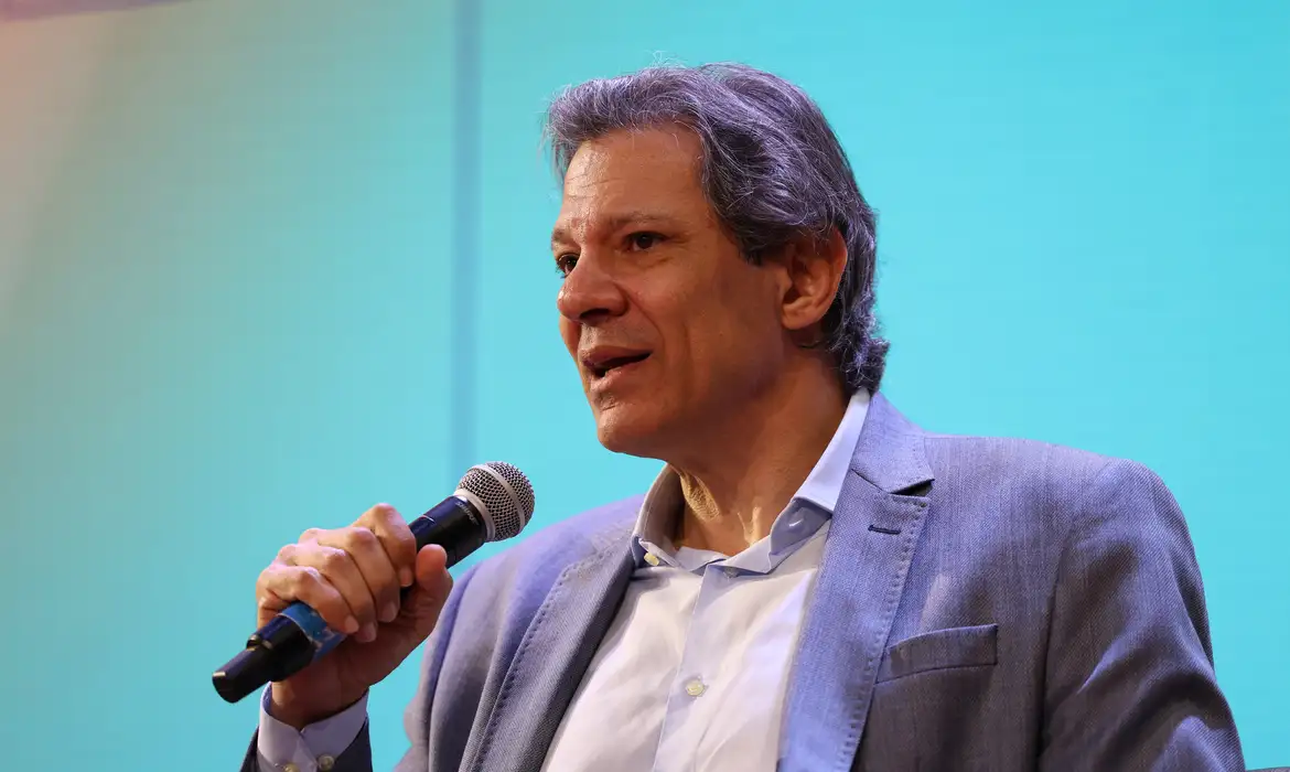 Ministro Fernando Haddad falando ao microfone em evento, com expressão de esclarecimento.