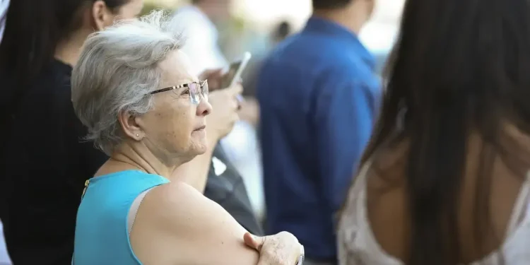 Idosa de cabelos curtos e óculos, vestindo blusa azul, observa o ambiente público ao seu redor.