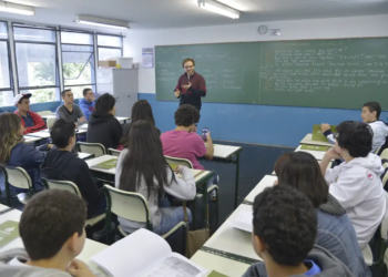 Sala de aula com alunos prestando atenção ao professor que está explicando na frente, escrevendo na lousa.