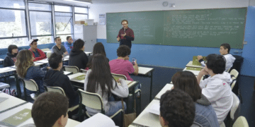 Sala de aula com alunos prestando atenção ao professor que está explicando na frente, escrevendo na lousa.