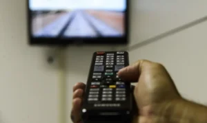 Pessoa segurando um controle remoto com uma TV ao fundo, representando a busca por canais digitais.