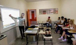 Estudantes em sala de aula assistindo aula no quadro branco, com o professor explicando.