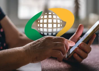 Idoso segurando um smartphone, com as mãos enrugadas, e o logo do INSS visível no fundo, com cores verde e amarela, simbolizando a liberação de pagamentos para aposentados.