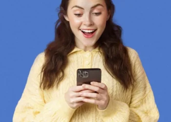 Pessoa sorrindo enquanto usa o celular com um fundo azul, mostrando expressão de surpresa ou empolgação."