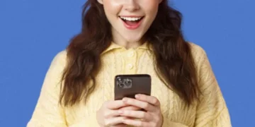 Pessoa sorrindo enquanto usa o celular com um fundo azul, mostrando expressão de surpresa ou empolgação."
