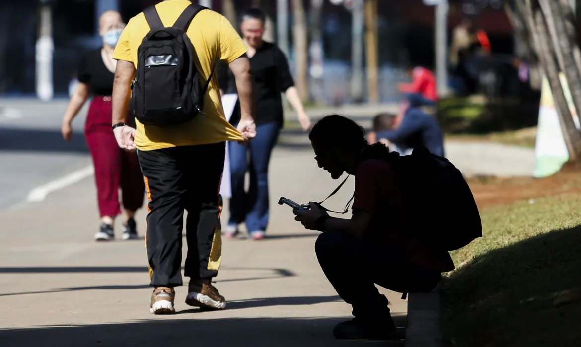 Estudantes caminhando na rua, com um jovem usando celular, representando os beneficiários do programa Pé-de-Meia.