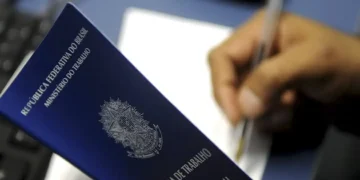 Carteira de Trabalho sendo segurada ao lado de uma pessoa assinando documentos.