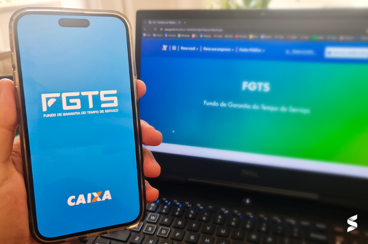 Pessoa segurando um celular com o aplicativo do FGTS aberto, enquanto um notebook exibe o site do FGTS ao fundo. A Mudança no Saque-Aniversário do FGTS de março de 2025 entra em vigor hoje, 13 de março.