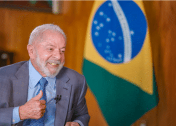 Presidente do Brasil em entrevista, sorrindo e fazendo um gesto positivo com a mão, com a bandeira brasileira ao fundo.