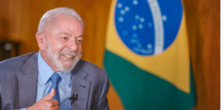 Presidente do Brasil em entrevista, sorrindo e fazendo um gesto positivo com a mão, com a bandeira brasileira ao fundo.
