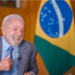 Presidente do Brasil em entrevista, sorrindo e fazendo um gesto positivo com a mão, com a bandeira brasileira ao fundo.