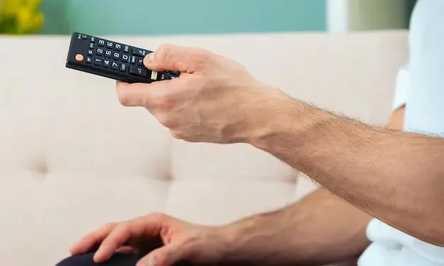 Pessoa segurando um controle remoto enquanto assiste TV no sofá.