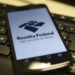 "Logo da Receita Federal no celular, com foco no aplicativo Meu Imposto de Renda, em fundo de teclado de computador."