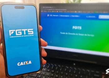 Aplicativo FGTS aberto no celular com a tela inicial destacando o logo da Caixa, e site oficial do FGTS ao fundo em um notebook