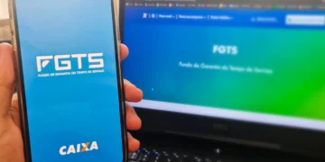 Aplicativo FGTS aberto no celular com a tela inicial destacando o logo da Caixa, e site oficial do FGTS ao fundo em um notebook