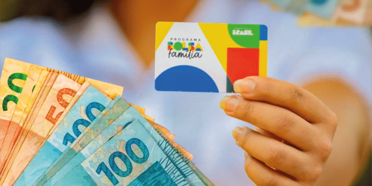 Imagem do cartão do Bolsa Família sendo segurado por uma mão, com cédulas de R$50 e R$100 ao fundo.