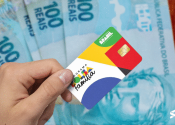 Pessoa segurando o novo cartão do Bolsa Família com notas de cem reais ao fundo
