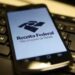 Logo da Receita Federal exibido em um smartphone com teclado ao fundo.