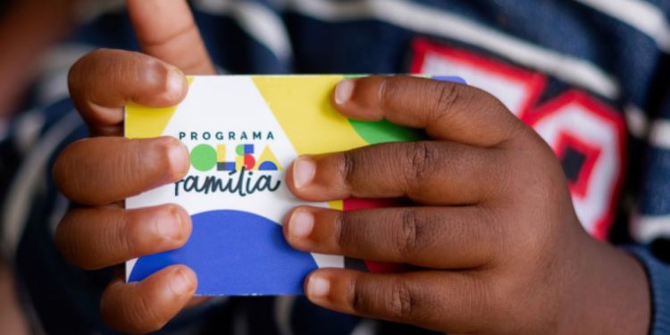 Cartão do programa Bolsa Família sendo segurado por uma criança, simbolizando a liberação do pagamento antecipado para 659 municípios.