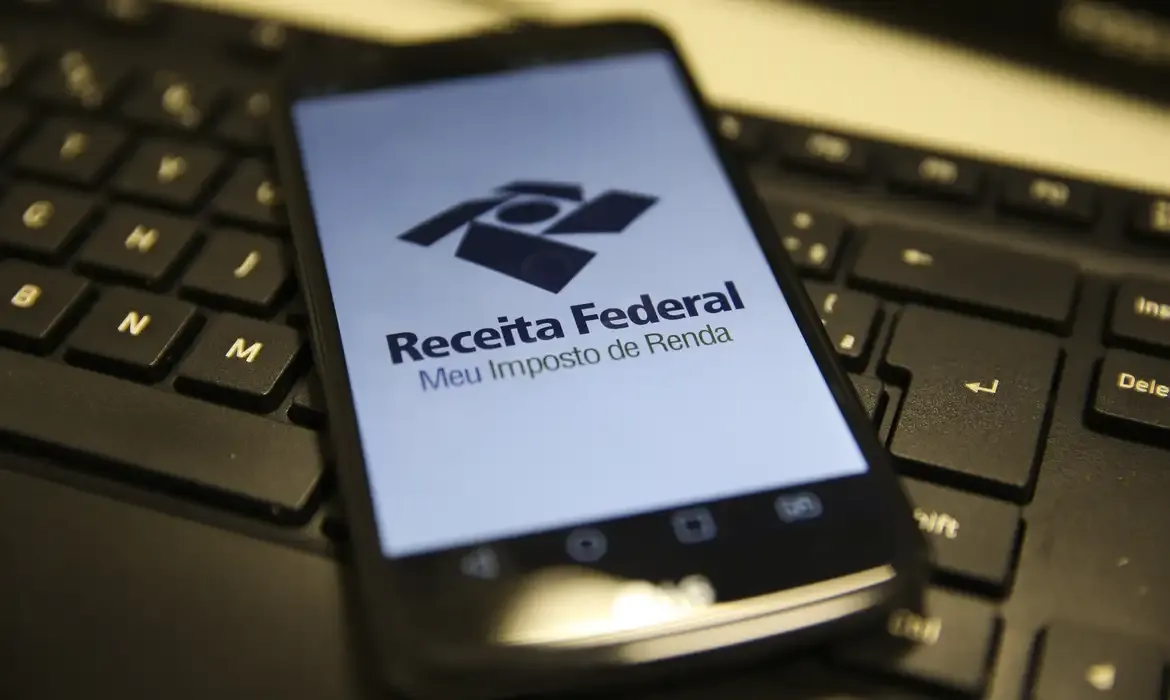 Smartphone exibindo o logo da Receita Federal e a frase 'Meu Imposto de Renda', com teclado de computador ao fundo.