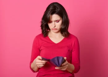 Mulher jovem olhando carteira vazia com expressão de preocupação.