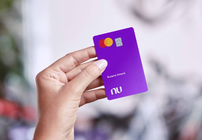 Pessoa segurando um cartão roxo do Nubank com o nome 'Suzana Amaral', relacionado à indenização de até R$ 10 mil.