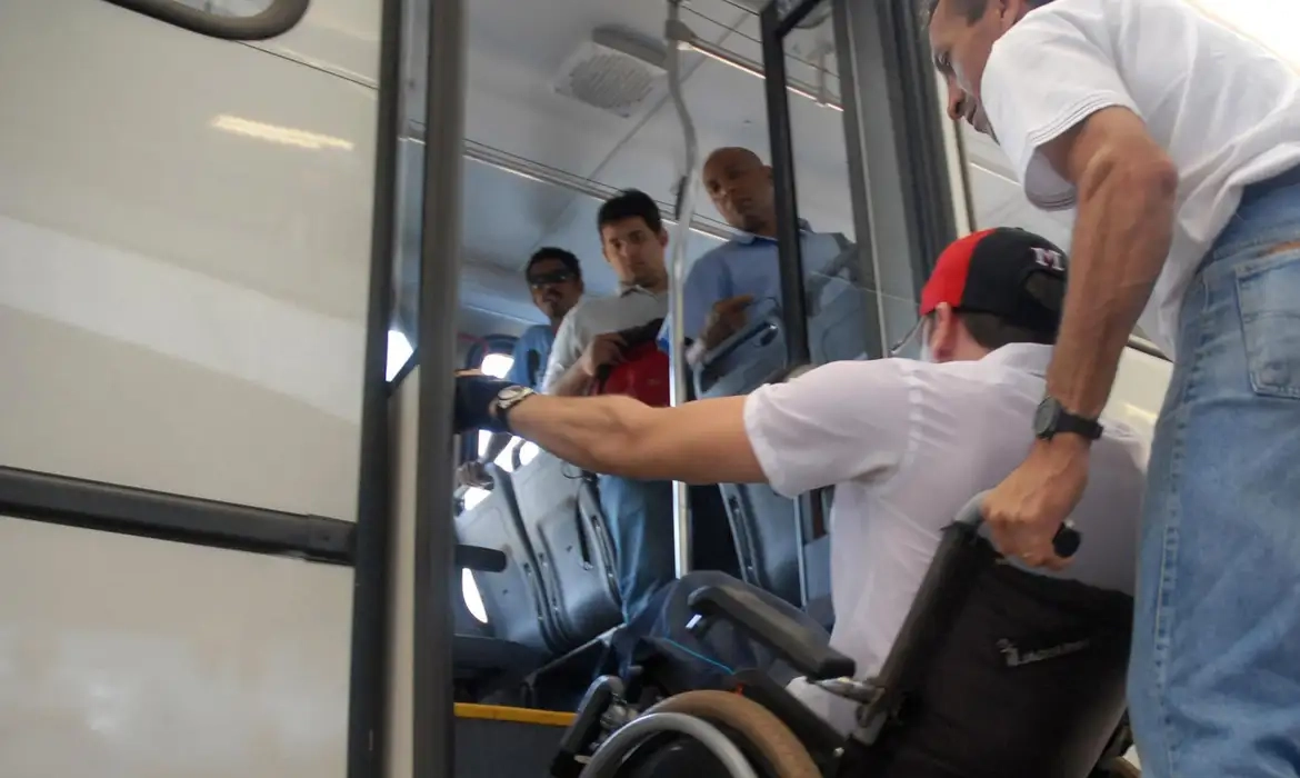 Homem em cadeira de rodas tentando embarcar em transporte público, auxiliado por outra pessoa, enquanto três homens observam.