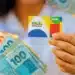 Cartão do Programa Bolsa Família sendo segurado por uma pessoa, com cédulas de dinheiro em destaque, representando o auxílio financeiro