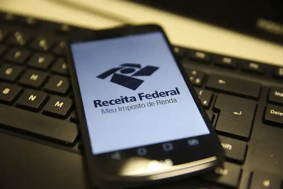 Smartphone exibindo o aplicativo da Receita Federal para consulta da restituição do Imposto de Renda sobre teclado de computador.