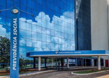 Fachada do prédio da Previdência Social com reflexo do céu azul nas janelas de vidro.