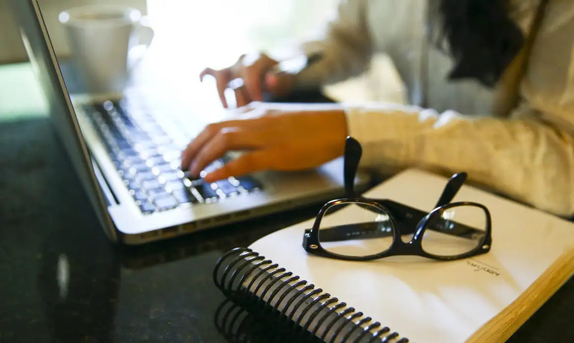 Pessoa digitando em laptop com óculos e caderno ao lado.