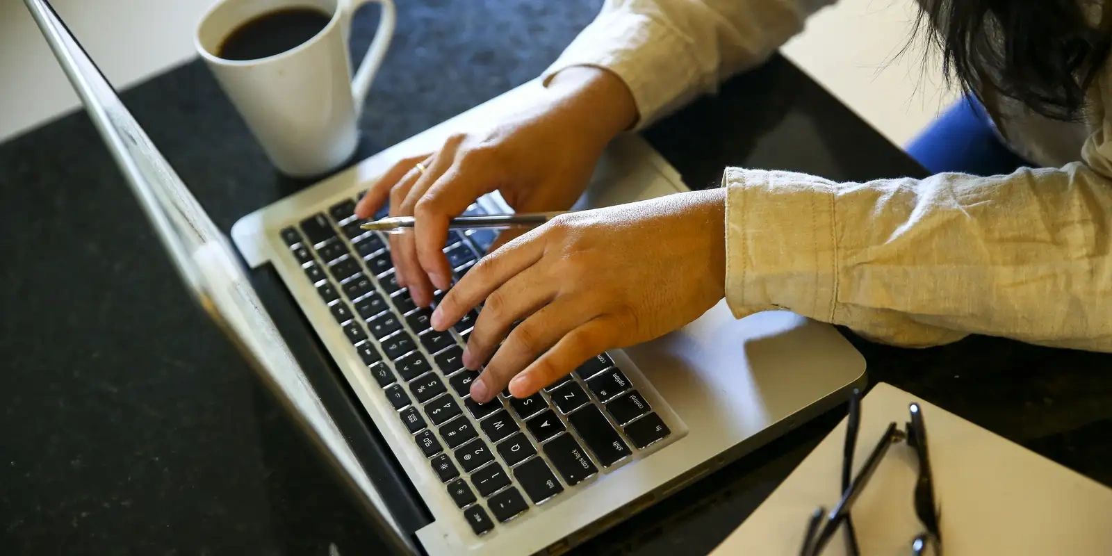 Pessoa usando laptop para se cadastrar em plataformas online, com café e óculos ao lado.