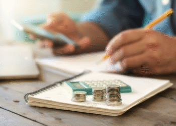 Pilha de moedas sobre caderno com calculadora ao fundo, enquanto pessoa usa celular e faz anotações.